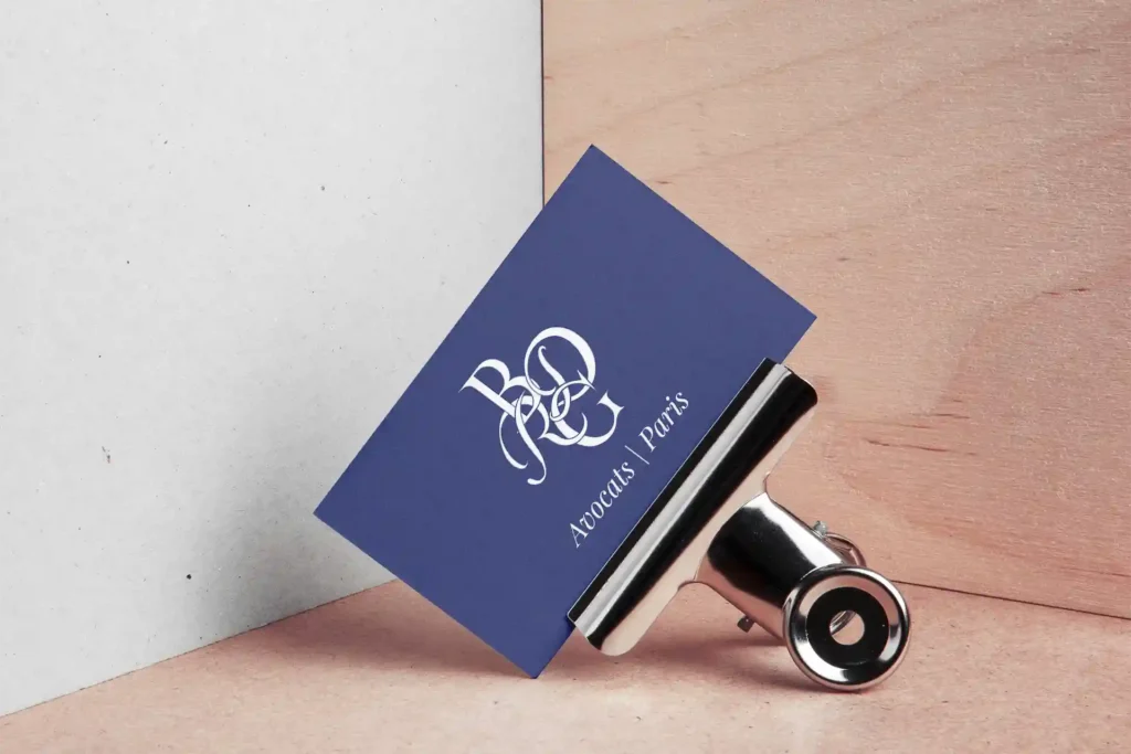 Carte de visite bleu marine du cabinet d'avocats BDRG Paris, monogramme calligraphié blanc, présentée sur fond bois et béton avec pince métallique