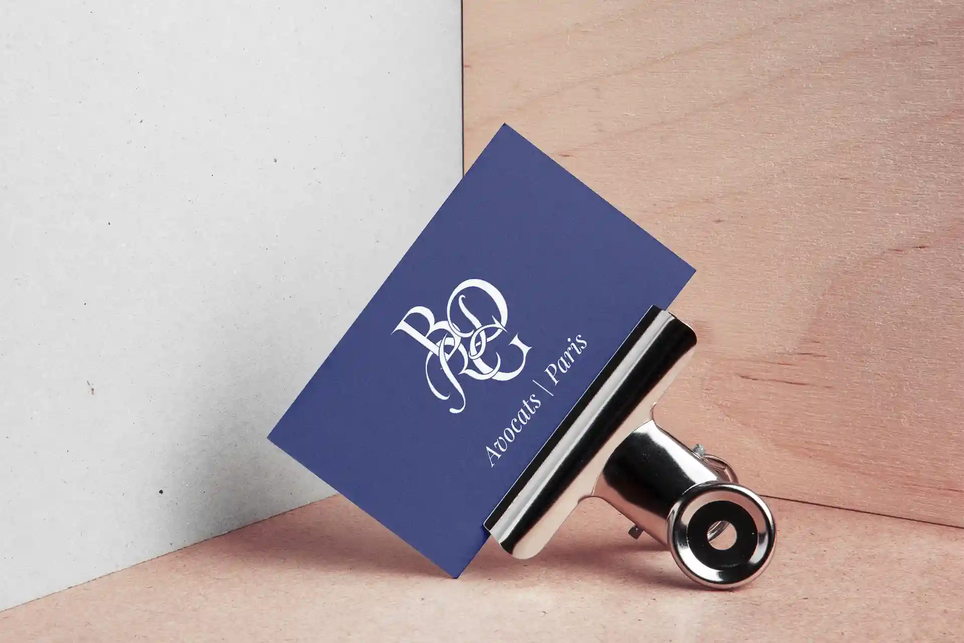 Carte de visite bleu marine du cabinet d'avocats BDRG Paris, monogramme calligraphié blanc, présentée sur fond bois et béton avec pince métallique